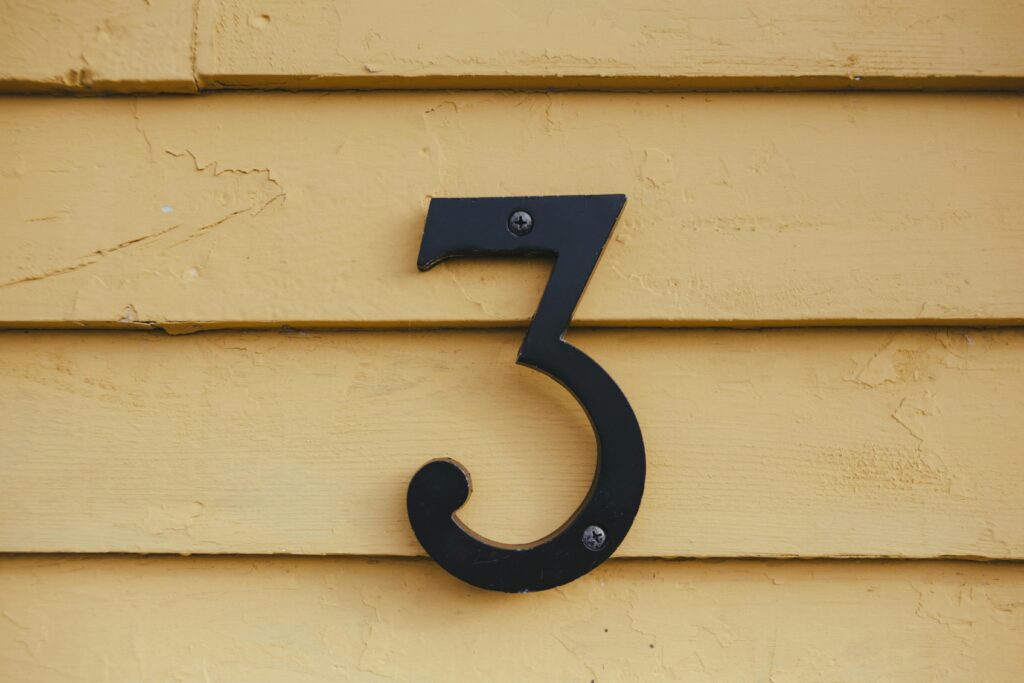 Close-up of a black number three sign mounted on a textured yellow wooden wall.